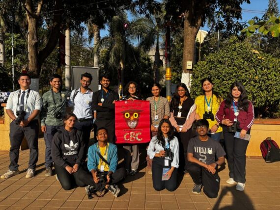 A group of students from Ninox Nature Club, Maharashtra, posing with binoculars and cameras during the Campus Bird Count 2025. They hold a red banner with an owl design and "CBC" written on it. The background features greenery and a sunny outdoor setting.