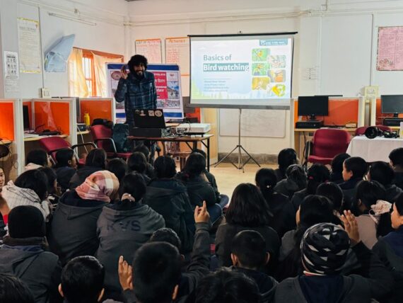 A presenter giving a presentation on "Basics of Birdwatching" to a classroom of students at Kendriya Vidyalaya School in Dirang, Arunachal Pradesh. The presenter stands next to a projection screen while students sit and raise their hands to participate.