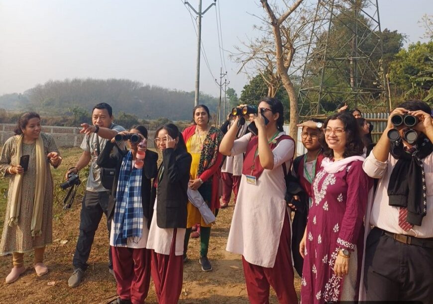 A group of women and men participating in a birdwatching session in Debendrachandranagar, Tripura, India. Some are holding binoculars and cameras, while others are observing their surroundings. The group is diverse, with participants in traditional attire and casual clothing. A power line and dry tree are visible in the background.