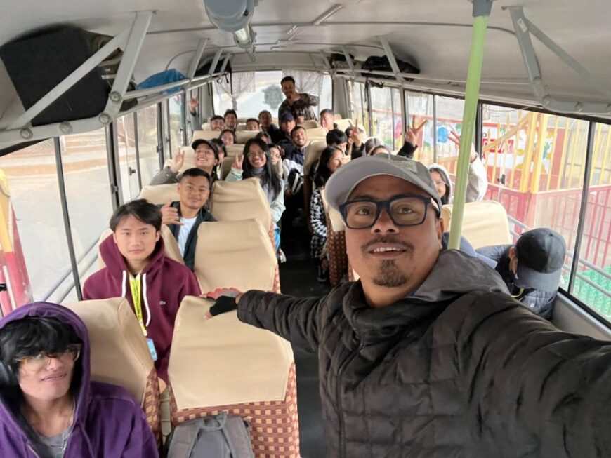 A group of cheerful birders inside a bus, smiling and posing for a selfie, taken by a man in a grey cap and glasses.