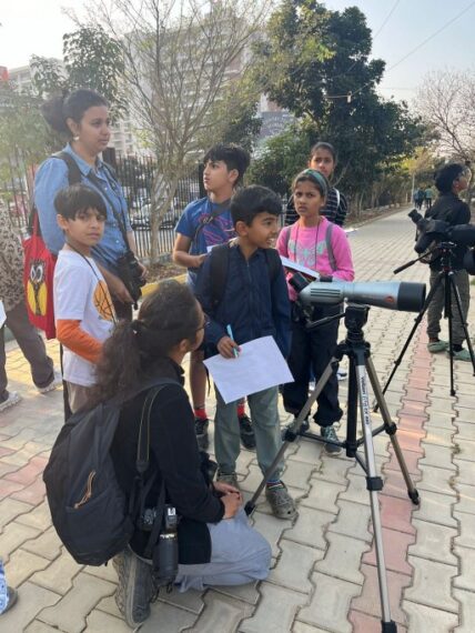 Group of children and two adults engaged in an outdoor birdwatching activity with a spotting scope and binoculars.