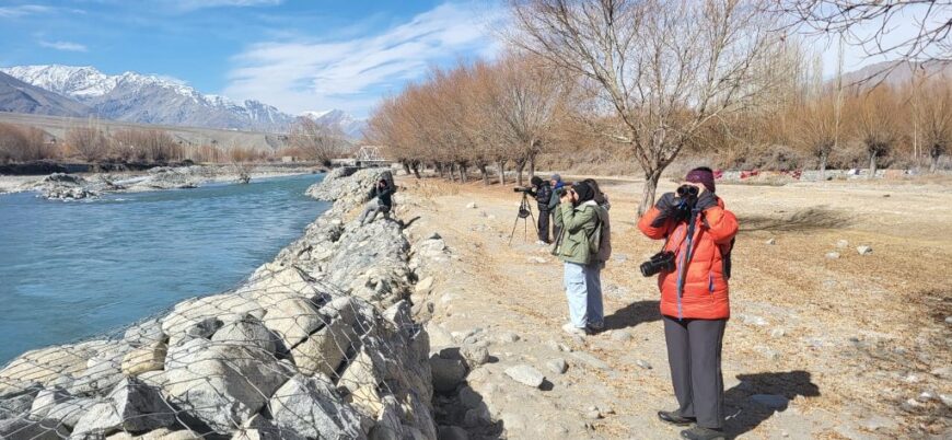 A group of birdwatchers observing birds along a rocky riverbank with snow-capped mountains in the background.