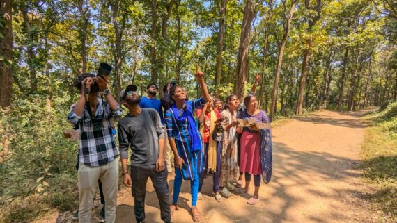 A group of people birdwatching in a forest trail, some pointing upward and others using binoculars and cameras.