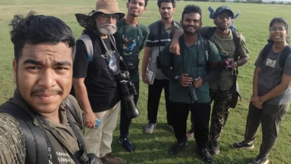 Group photo of seven birdwatchers from Oorna Nature Club at NISER, Bhubaneswar, taken during the Rajo Bird Count 2025. Some participants are holding cameras and binoculars, standing on an open grassy field.