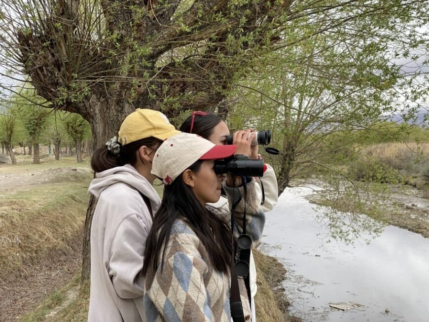Three young women birding with binoculars along a stream in Ladakh, surrounded by trees with fresh green leaves.